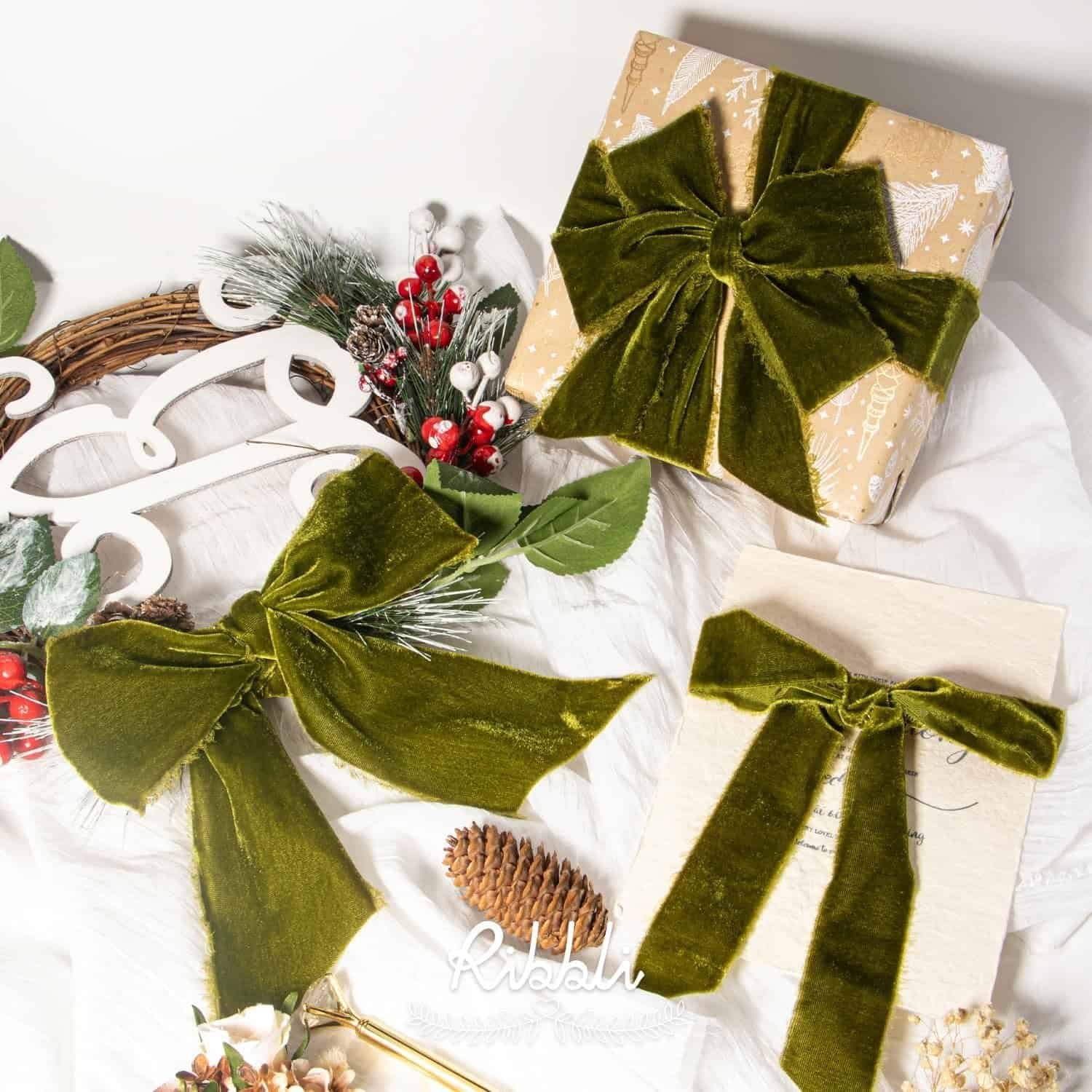 Green velvet bows decorate a wrapped gift and a card, with festive holiday decorations and greenery displayed on a white surface.