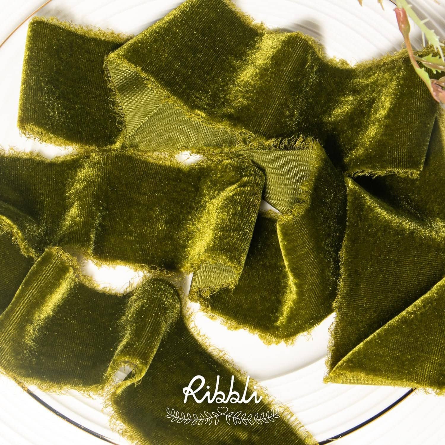 Olive green velvet ribbon with frayed edges arranged loosely on a white plate, with a small plant visible at the edge. The word "ribbli" appears at the bottom.