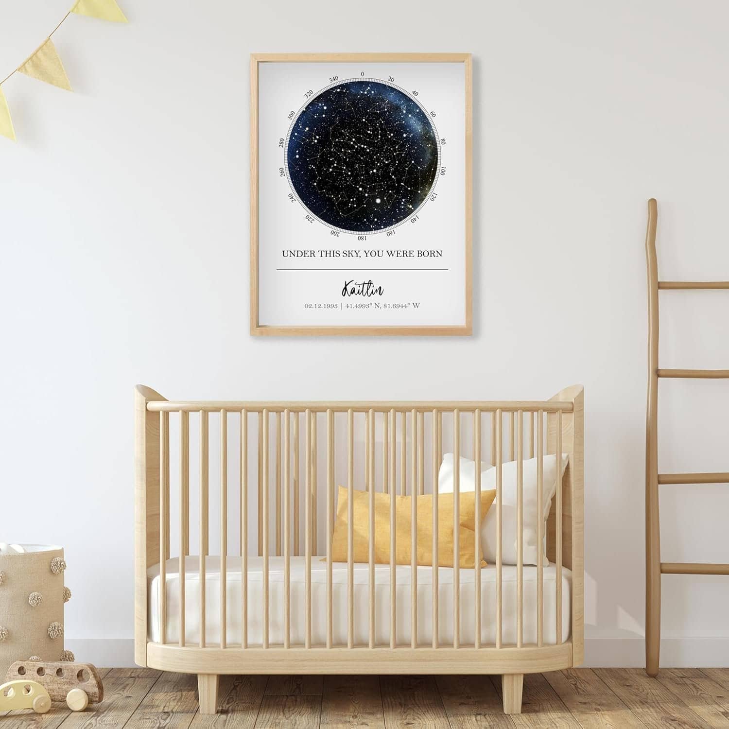 A minimalist nursery with a wooden crib, yellow and white pillows, wall art above the crib showing a star map, and simple decor including a ladder and bunting.