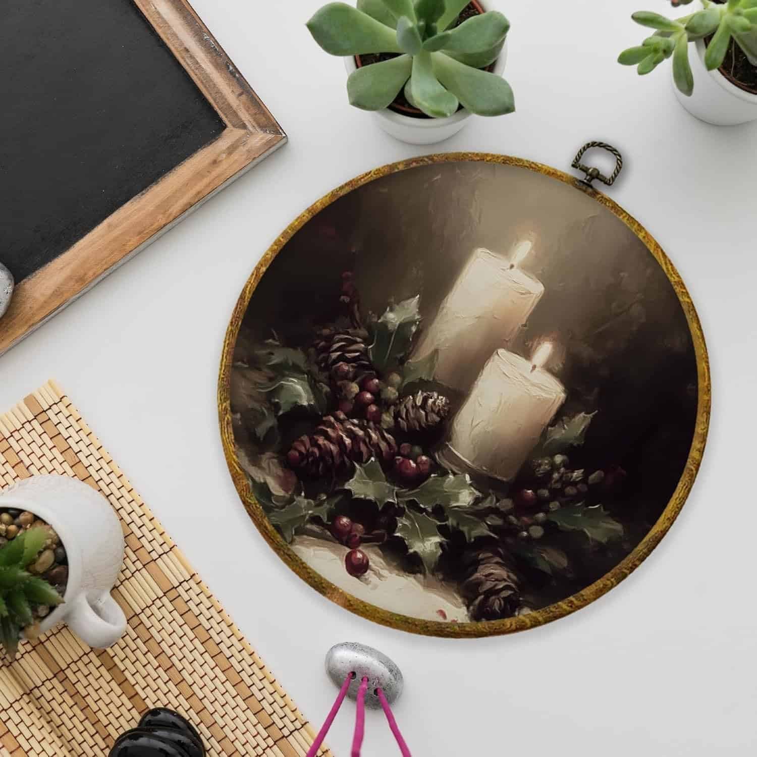 A round decorative plate featuring two lit white candles, pinecones, holly leaves, and red berries, placed on a white tabletop near potted succulents and a chalkboard.