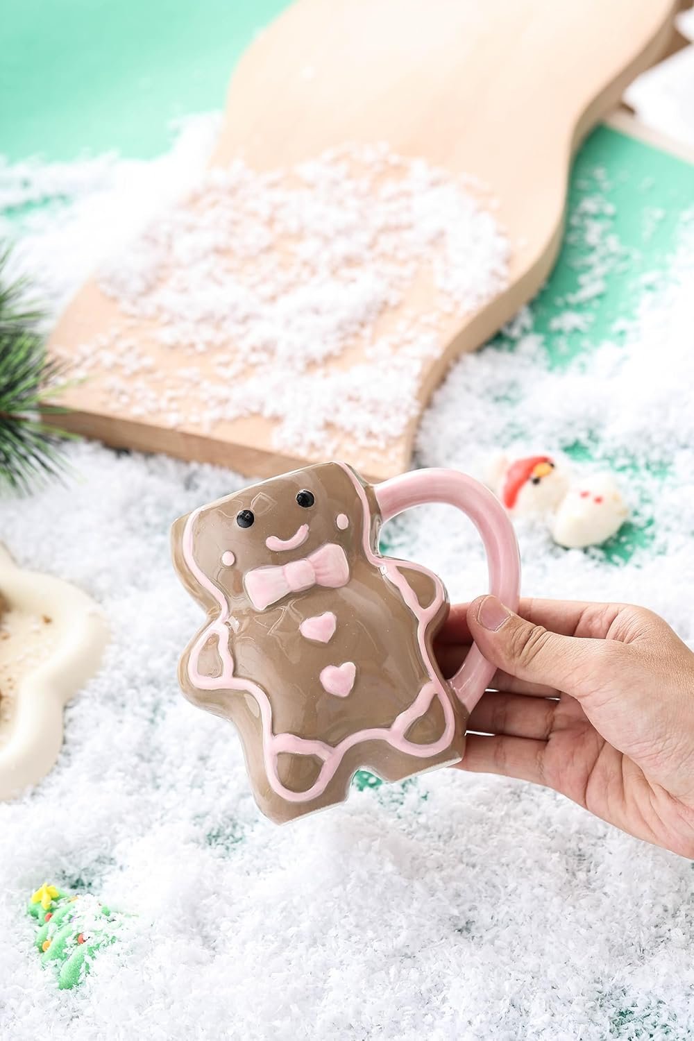 A hand holds a mug shaped like a smiling gingerbread person with pink accents, surrounded by artificial snow and festive decorations.