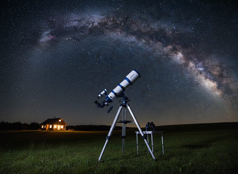 A large telescope on a tripod stands on grass at night, with a small lit house in the background and the Milky Way shining brightly in the star-filled sky above.