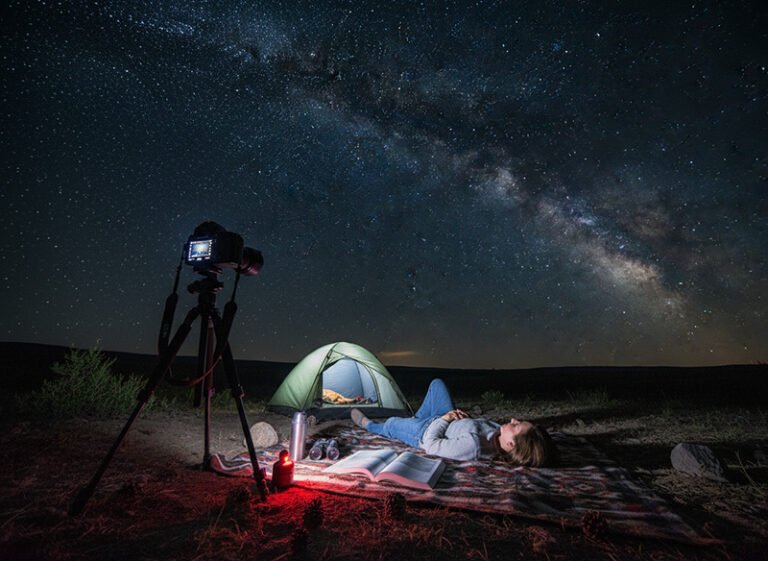 A person lies on a blanket near a tent and camera, stargazing under a clear night sky with the Milky Way visible, eagerly awaiting the Perseids meteor shower 2026.