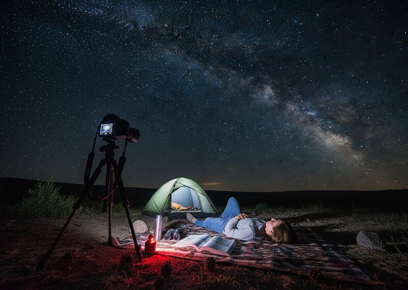 A person lies on a blanket near a tent and camera, stargazing under a clear night sky with the Milky Way visible, eagerly awaiting the Perseids meteor shower 2026.