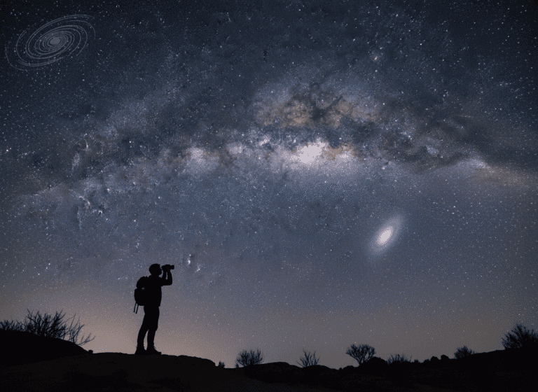 A silhouette of a person with a backpack looks at a star-filled night sky, marveling at the glowing galaxies overhead, while standing on uneven ground dotted with sparse bushes.