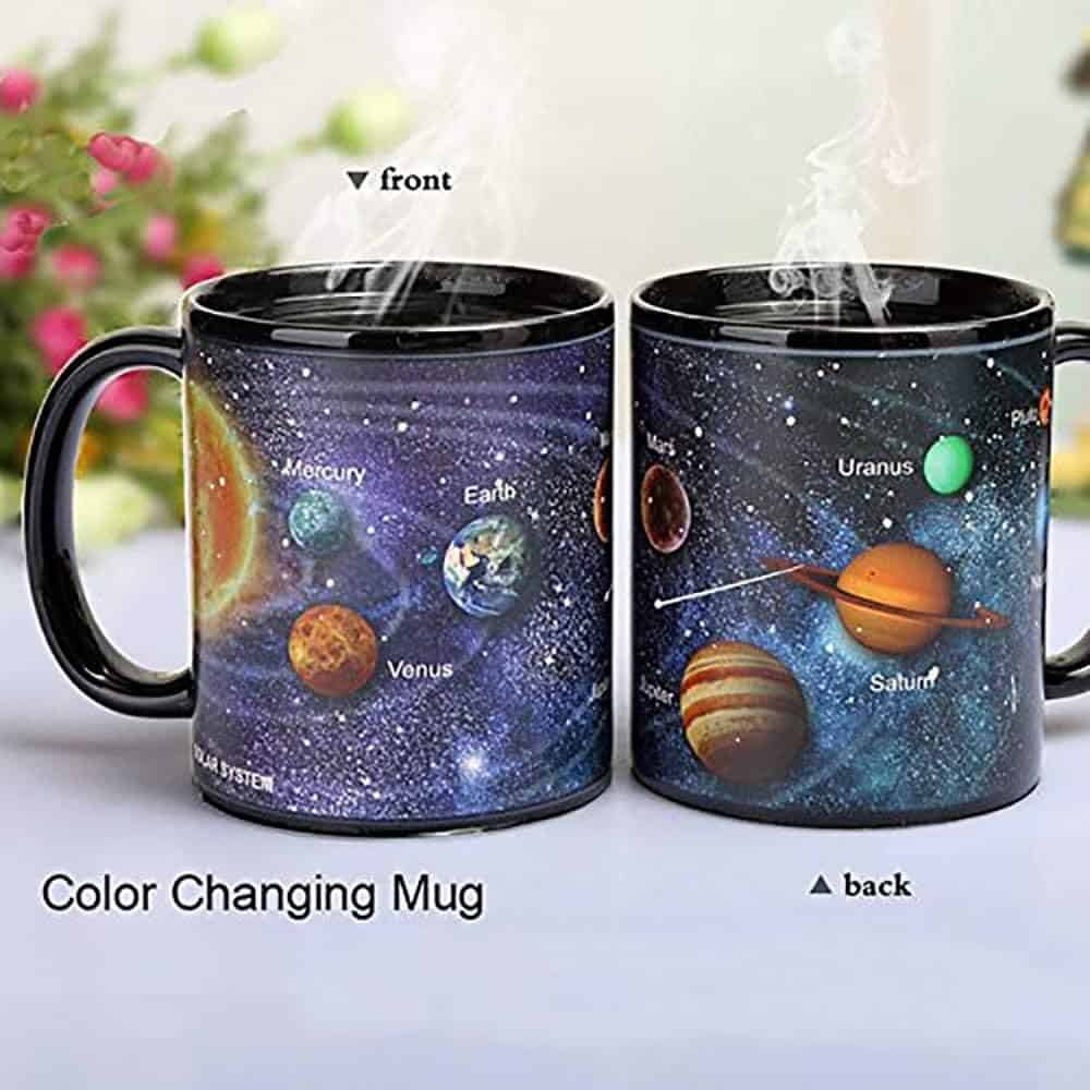 Two steaming color-changing mugs display a solar system design with labeled planets on a table; plants are blurred in the background.