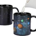 A hand pours hot water into a black mug, revealing a colorful solar system design with labeled planets. Another mug shows the plain black design with white stars.
