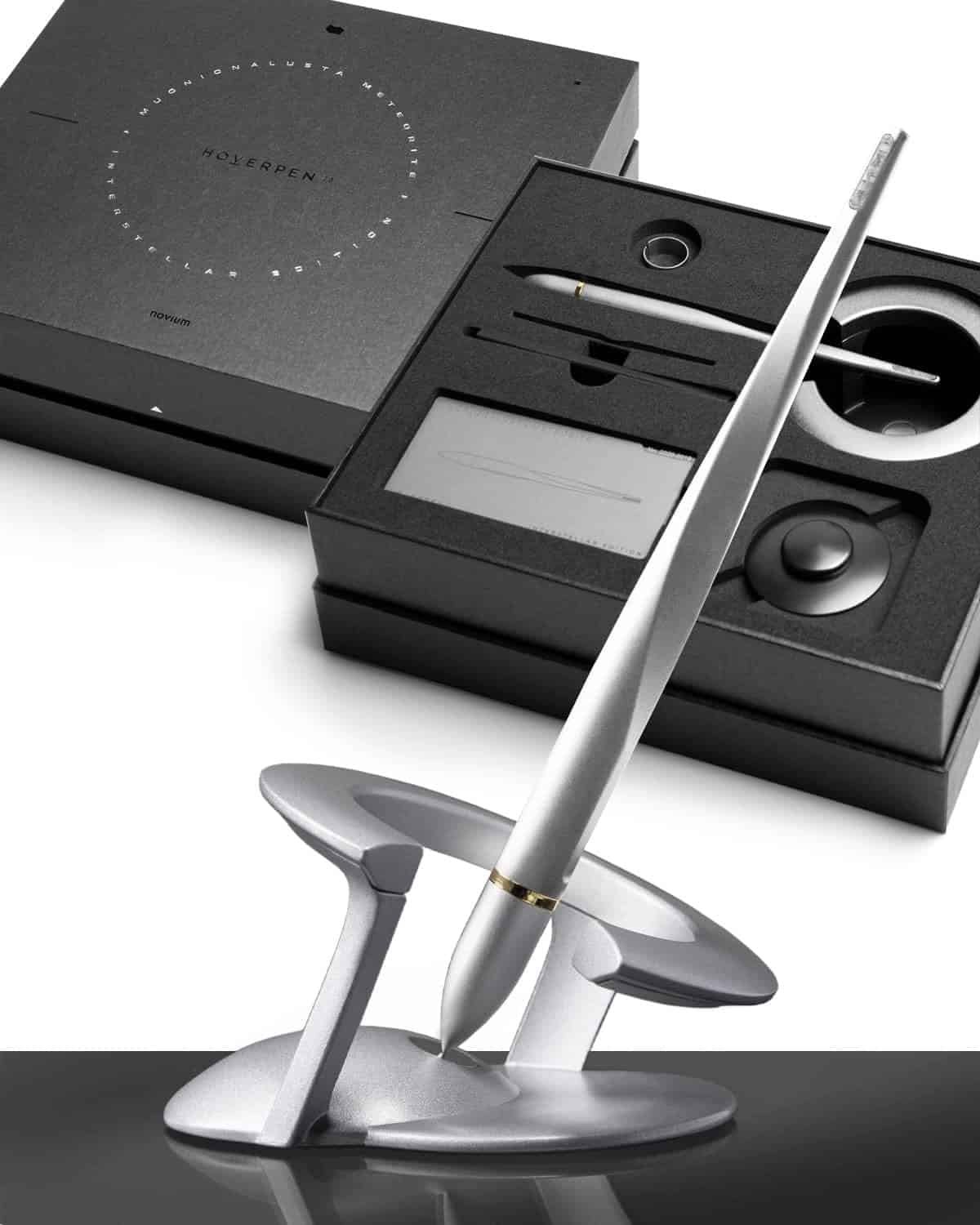 A sleek metallic pen is displayed on a modern stand, with an open black presentation box in the background containing additional pen accessories.