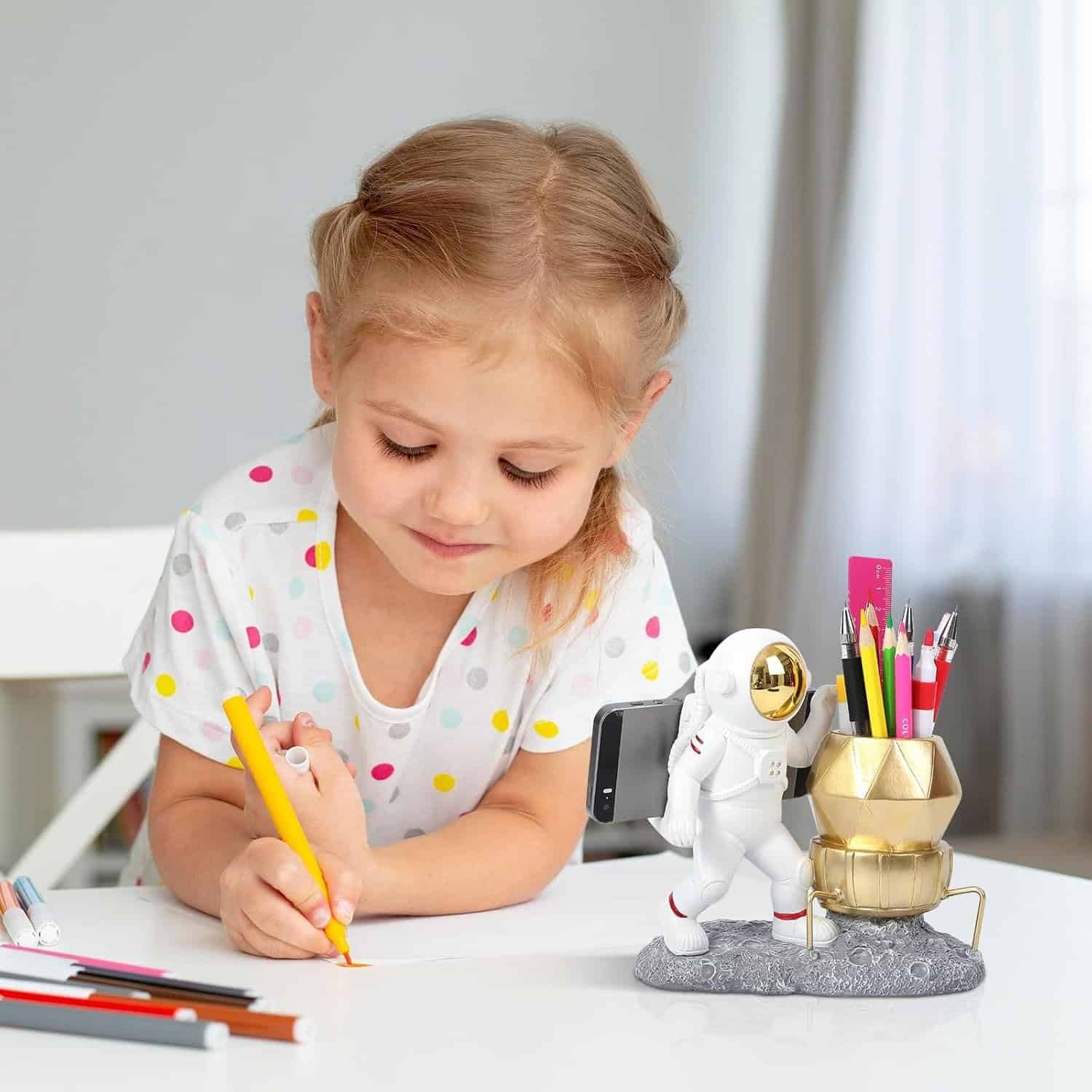 Young girl in a polka dot shirt drawing with colored pencils at a table, with a space-themed pen holder and phone stand beside her.