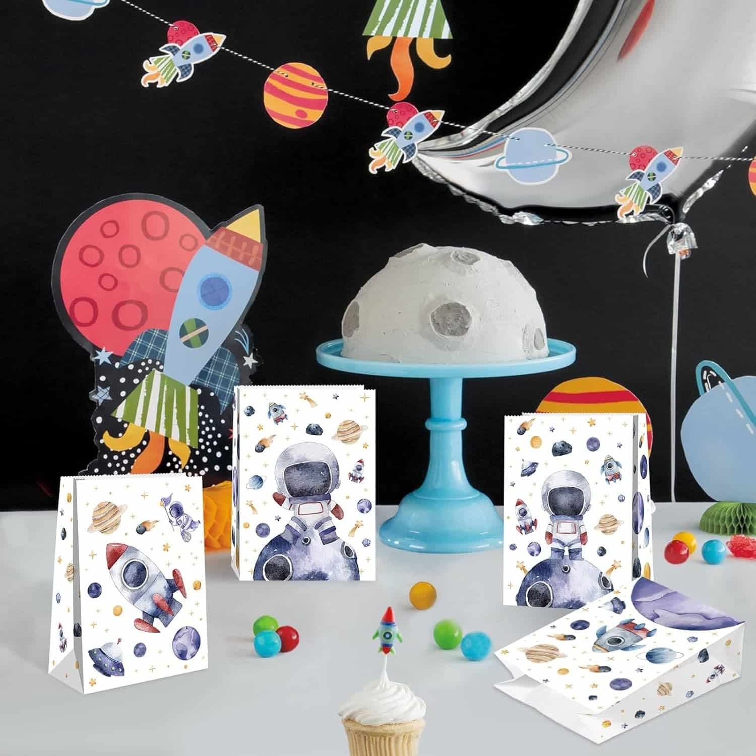 Space-themed party table with a moon cake on a blue stand, astronaut and rocket decorations, planet garland, goodie bags, colored candies, and a cupcake with a rocket topper.