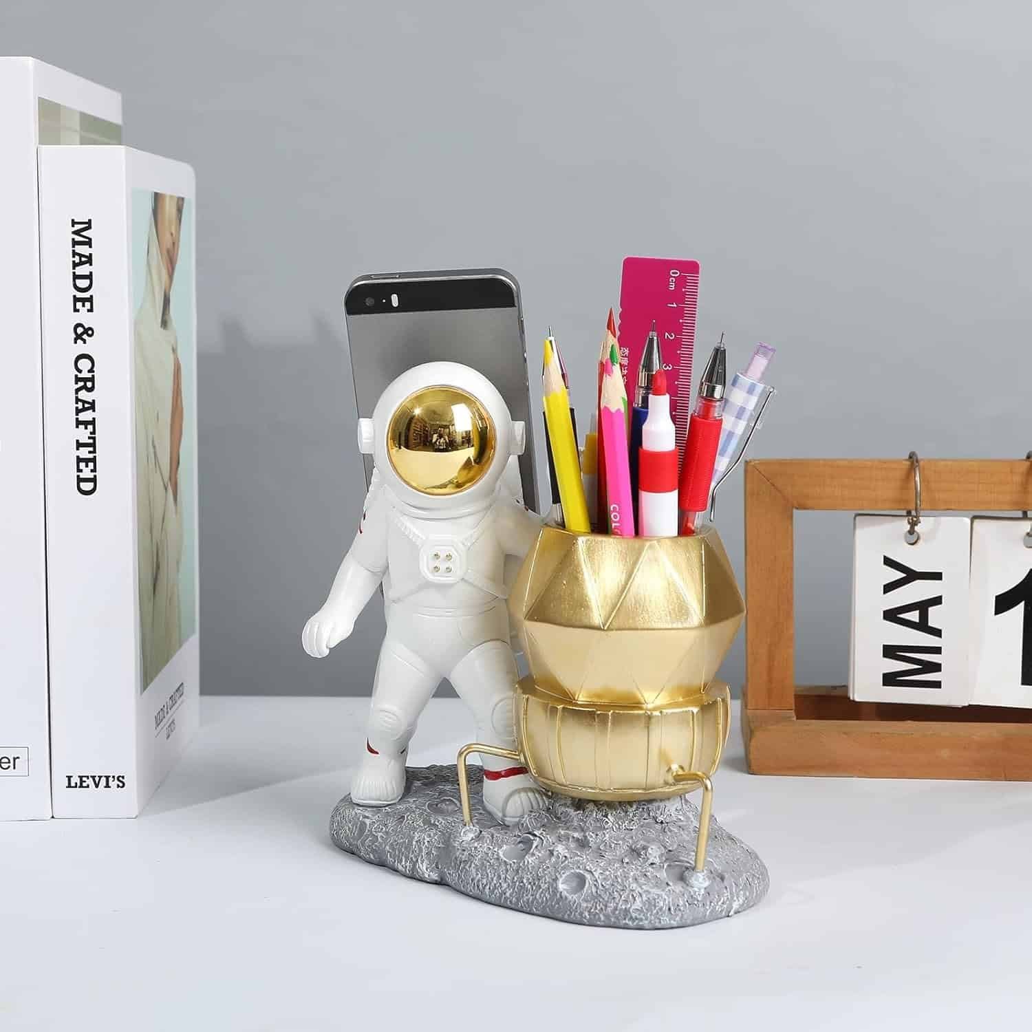 A desk organizer shaped like an astronaut holding a phone and a gold lunar module holding pens, next to a calendar displaying May 18 and books on a gray surface.