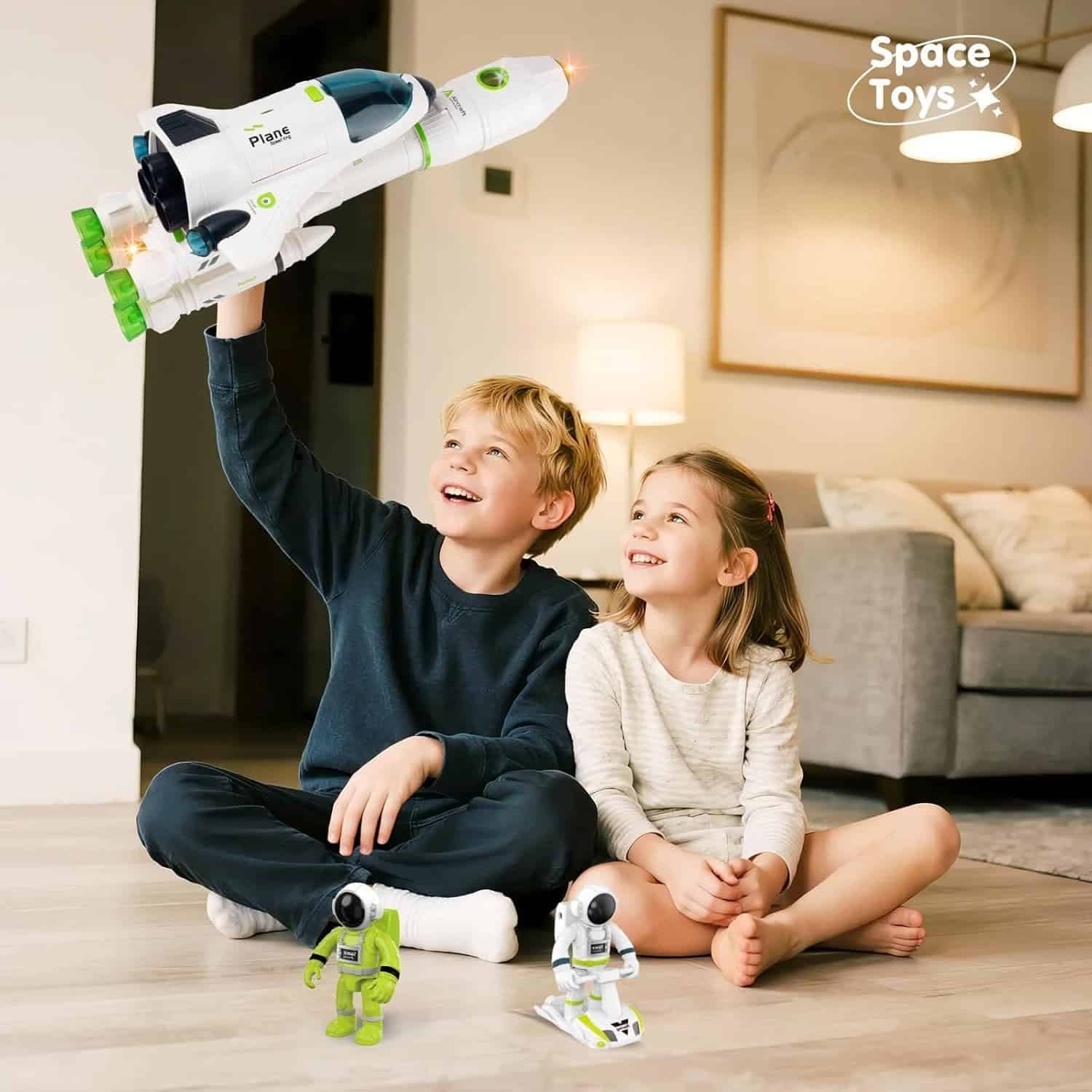 Two children sit on the floor indoors, playing with a toy spaceship and astronaut figures. The boy holds the spaceship up while the girl watches and smiles. “Space Toys” is written in the corner.