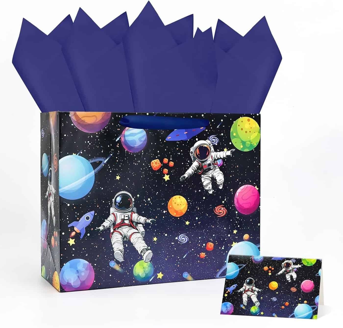 A gift bag with a space theme featuring astronauts, planets, and stars on a dark background, with dark blue tissue paper and a matching gift card.