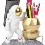 A desk organizer shaped like an astronaut and lunar lander, holding a smartphone and various pens, pencils, and a ruler on a gray, moon-like base.