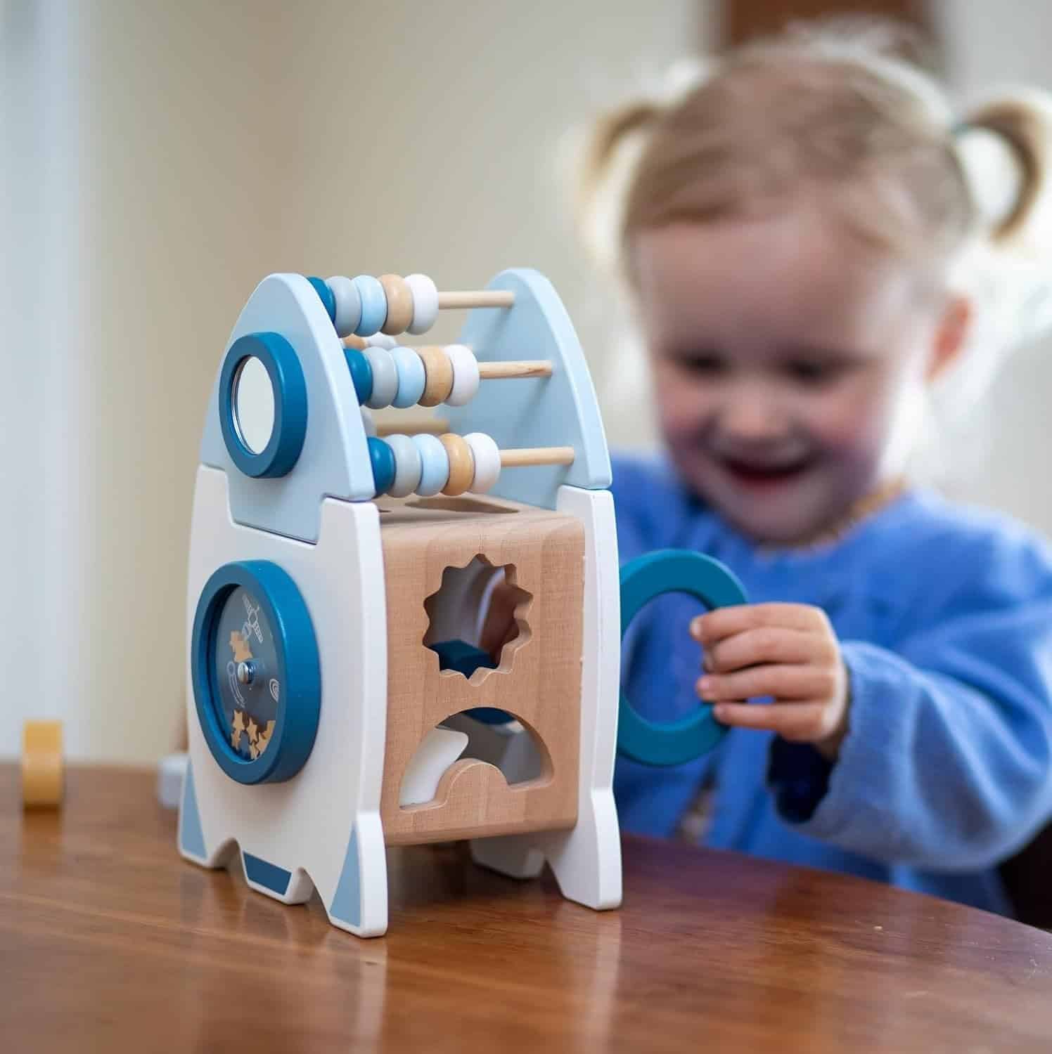 A young child plays with a wooden rocket-shaped activity toy featuring an abacus, shape sorter, and spinning gears on a wooden table.
