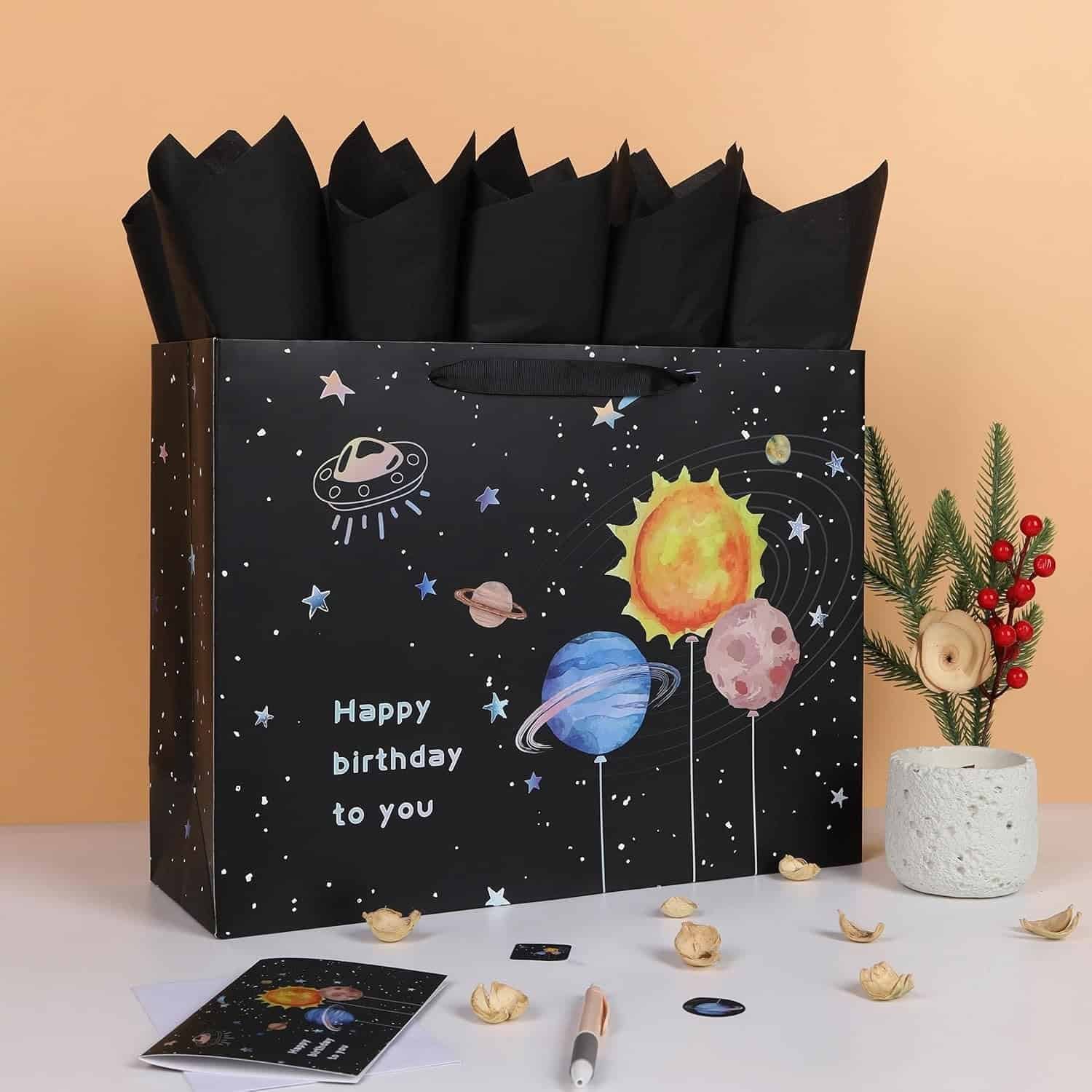 A large gift bag with a space and planets design, "Happy birthday to you" text, black tissue paper, and a small plant with red berries on a table.