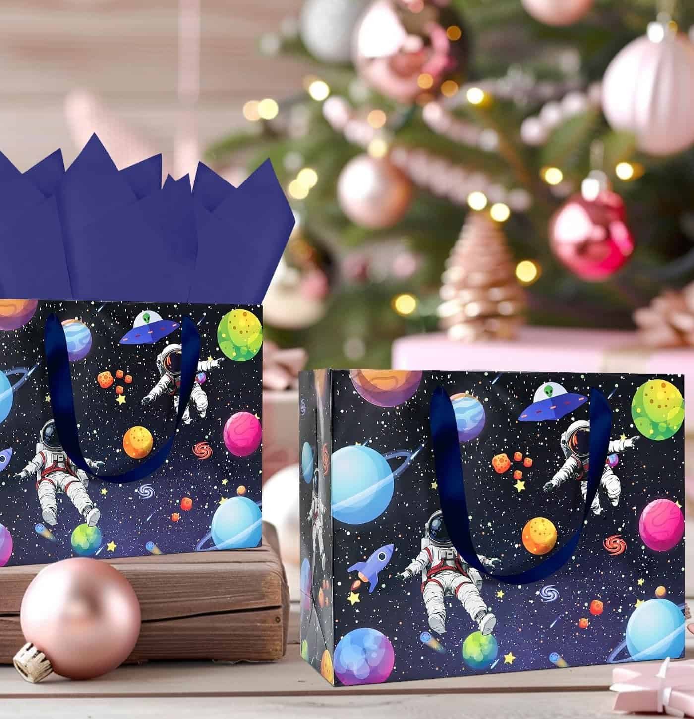 Two gift bags with space-themed designs featuring astronauts, planets, and UFOs are placed in front of a decorated Christmas tree with ornaments and wrapped gifts.