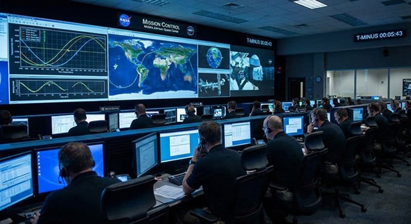 People at desks monitor computers in a NASA mission control room, where large screens display maps, graphs, and NASA spacecraft data at the front.