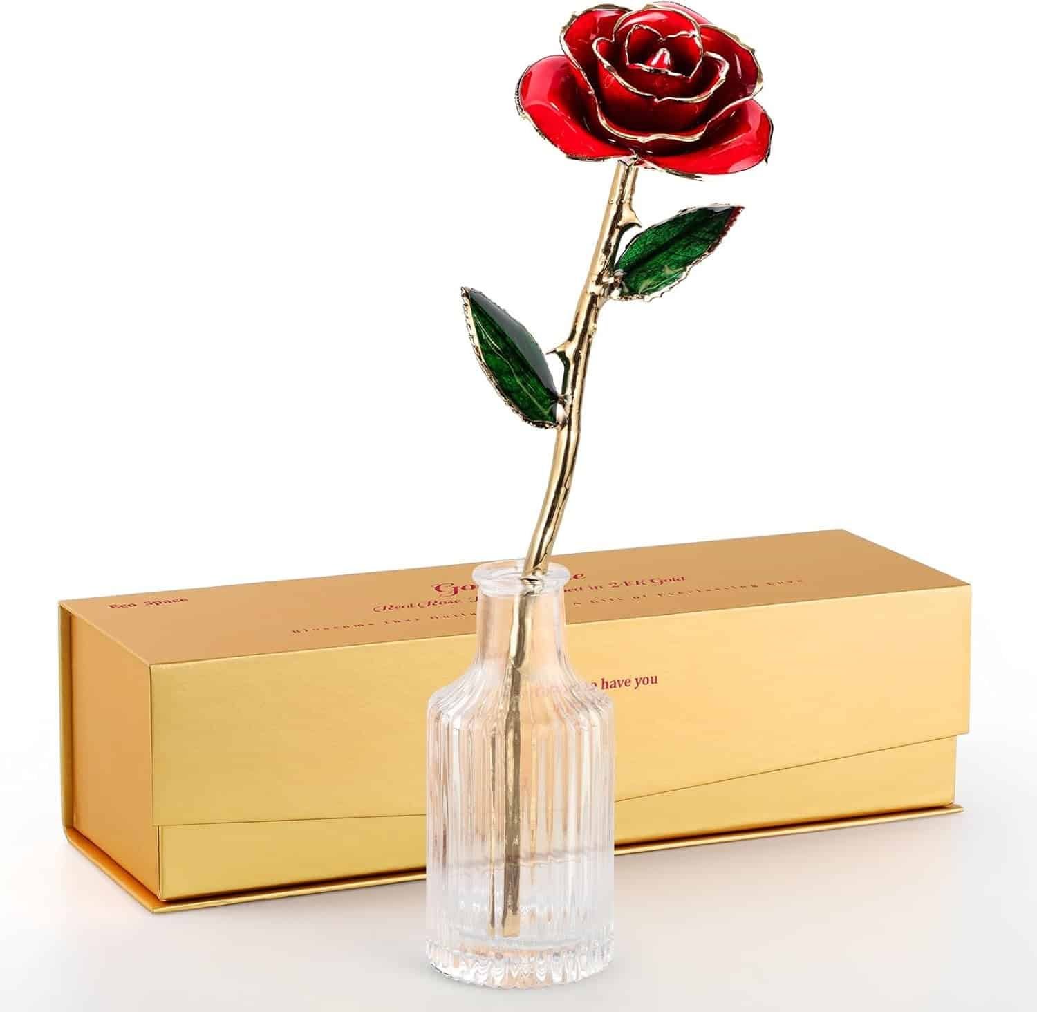 A single artificial red rose with a gold stem and green leaves in a glass vase, placed in front of a gold-colored gift box.
