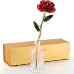 A red artificial rose with gold trim stands in a clear glass vase, positioned in front of a closed gold gift box.