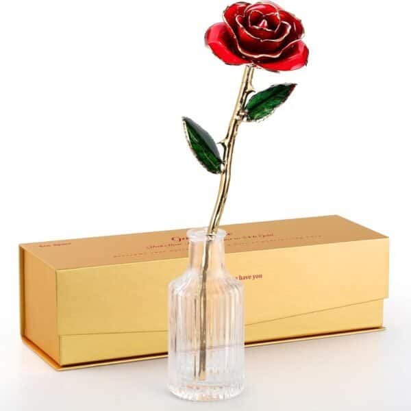 A red artificial rose with gold trim stands in a clear glass vase, positioned in front of a closed gold gift box.