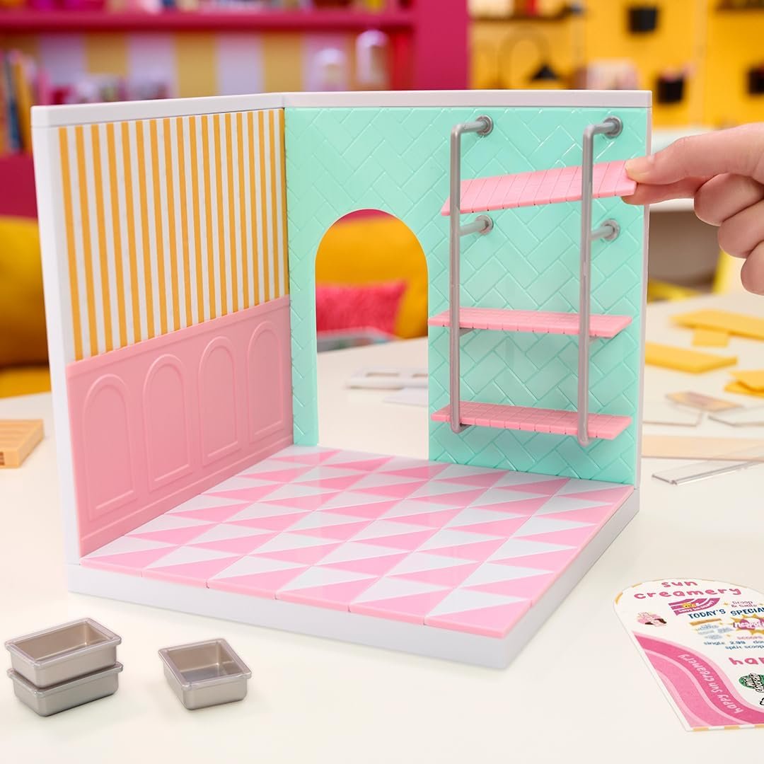 A hand assembles pink shelves inside a miniature pastel room with patterned floor and walls; small metal trays and paper cutouts are on the table.