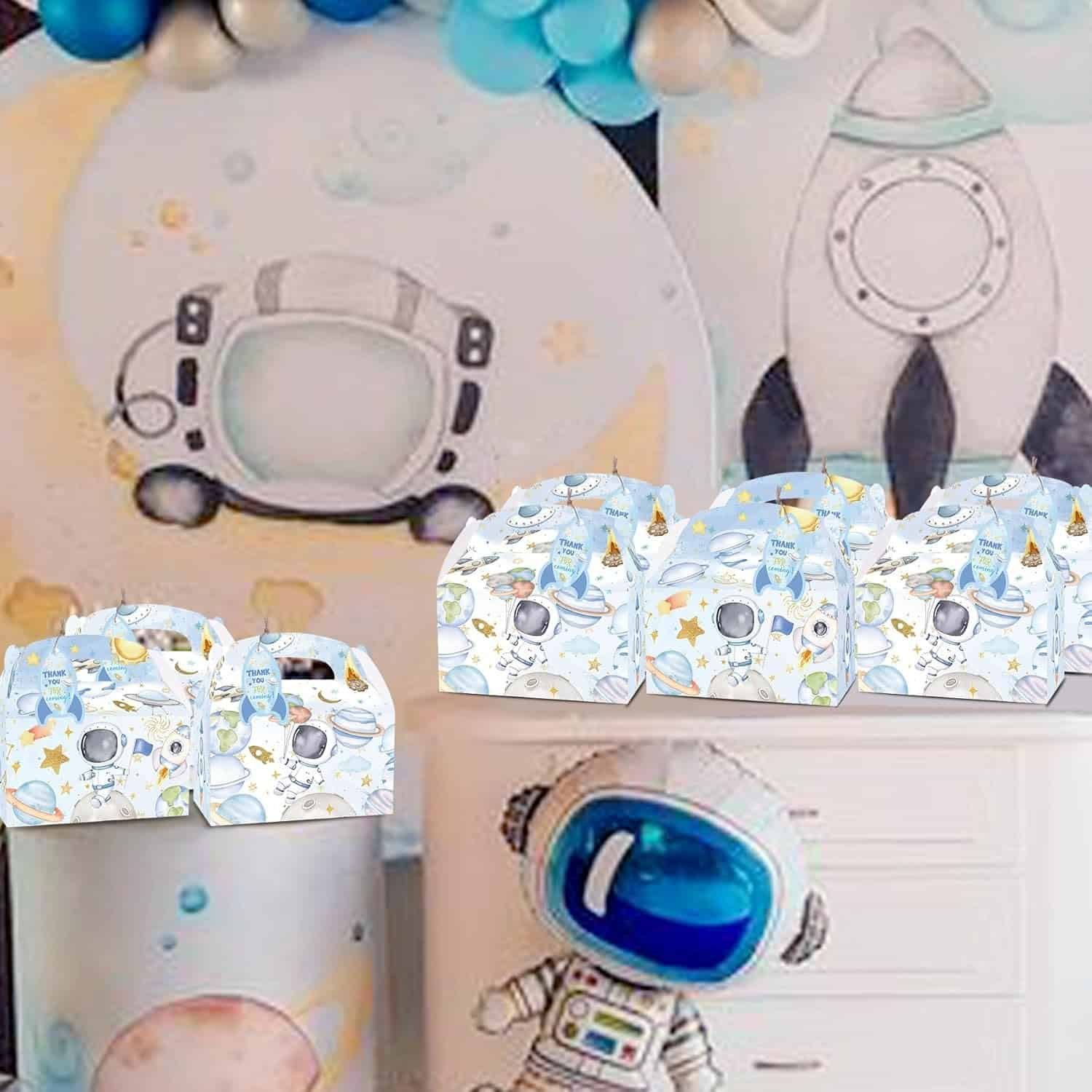 Space-themed party decorations with a rocket and astronaut illustrations, blue and white balloons, and matching favor boxes displayed on a table.