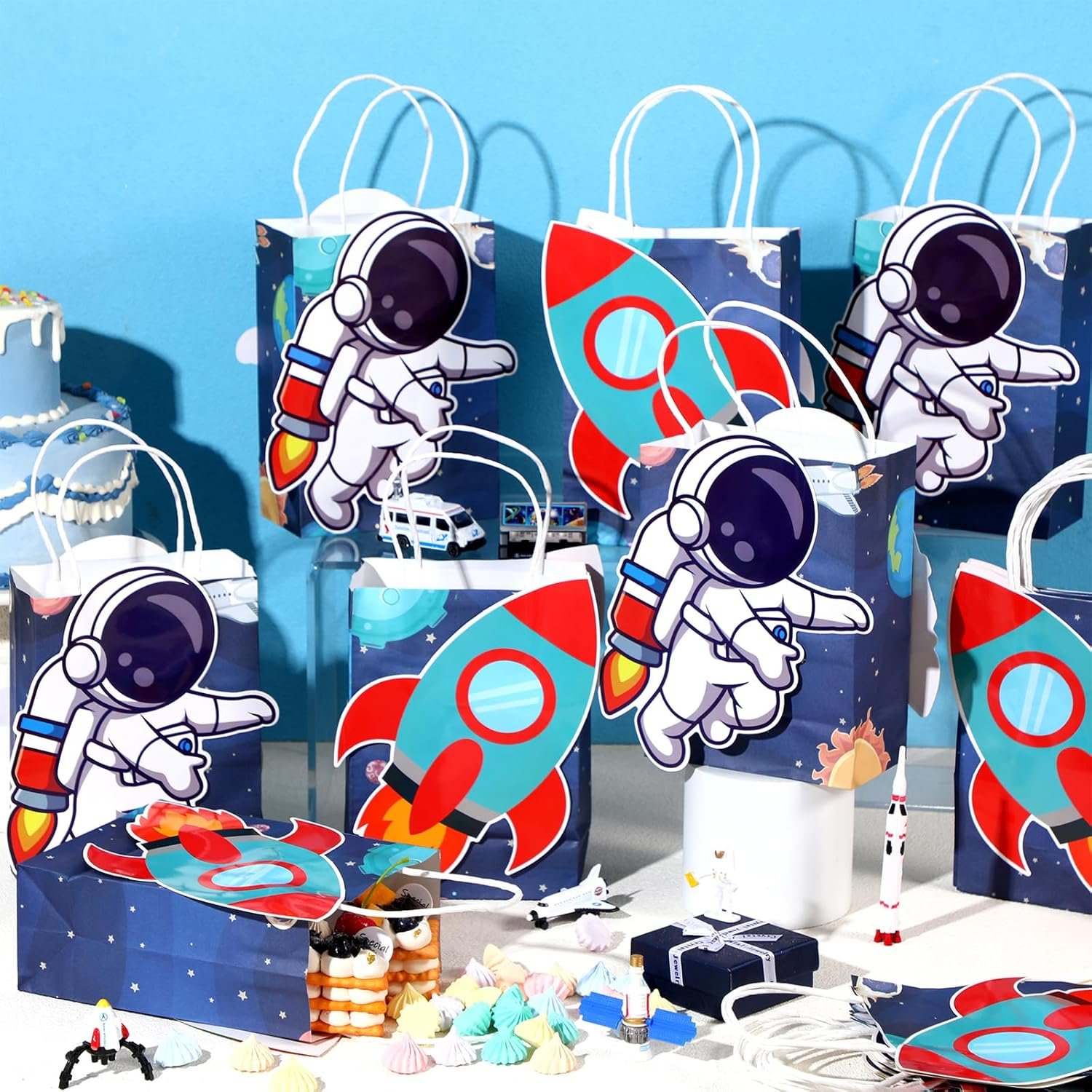 Gift bags with astronaut and rocket designs are arranged on a table with party treats, cake, miniature rockets, and a blue background.