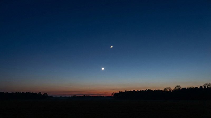 A twilight sky with a visible crescent moon and a bright planet above the horizon hints at a planetary alignment, with silhouettes of trees at the bottom.