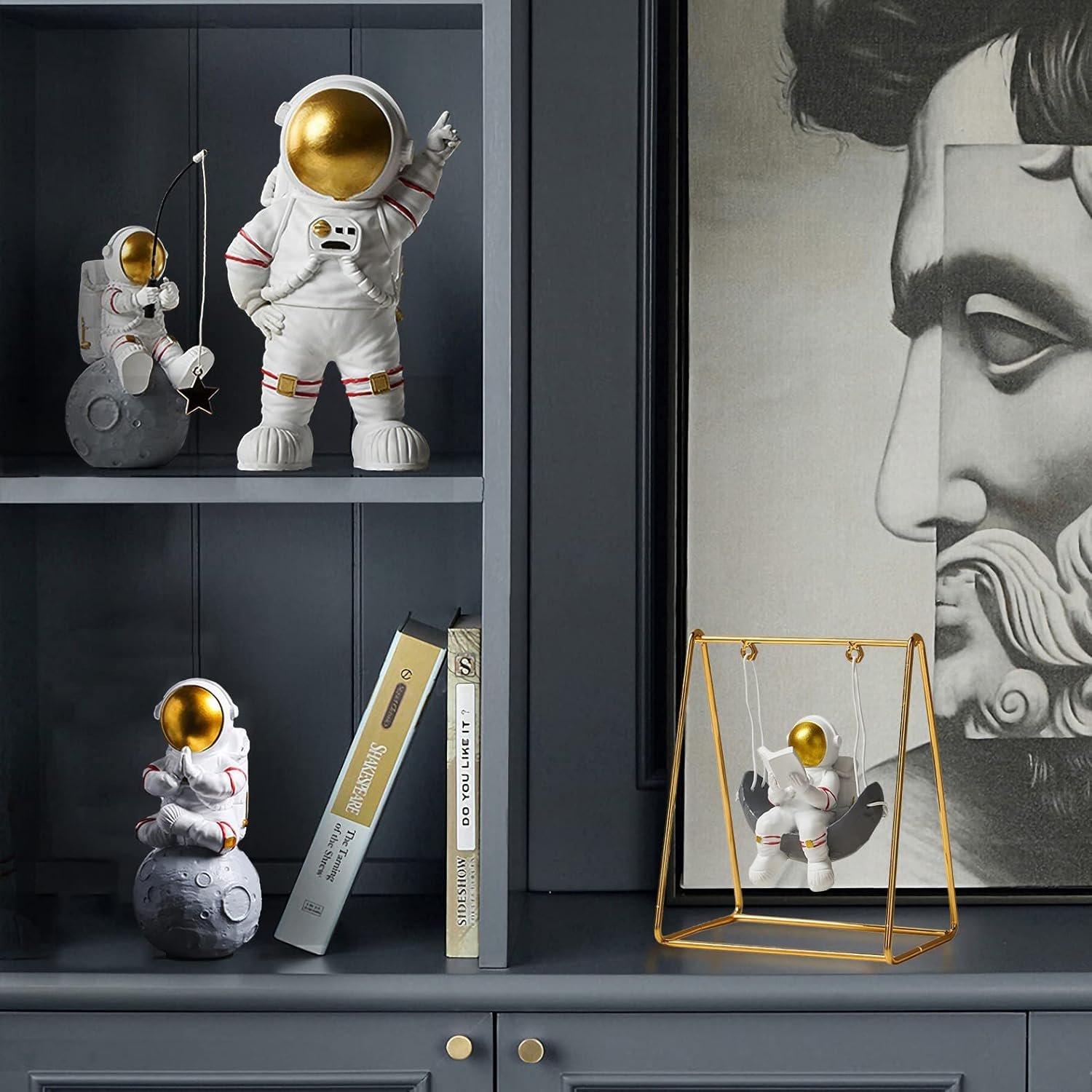 Three astronaut figurines are displayed on a gray shelf alongside books; one astronaut is on a swing, with part of a large monochrome portrait visible in the background.