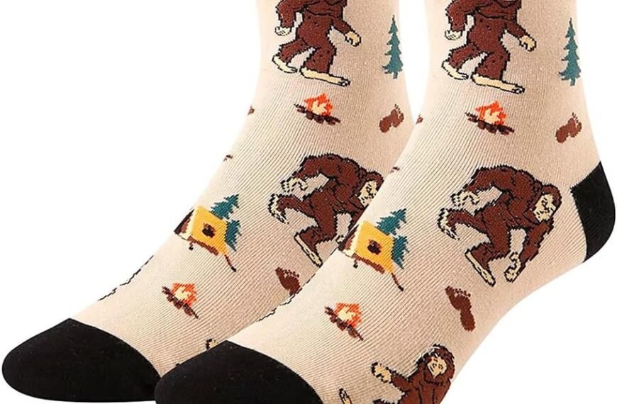 Pair of beige socks with black cuffs featuring cartoon Bigfoot, tents, campfires, and trees; shown with a polka-dot gift box and a view of the socks folded inside.