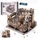 A wooden mechanical marble run model kit called "Marble Spaceport" is shown assembled, with gears, tracks, marbles, and its packaging box in the background.