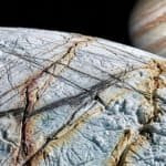 A close-up view of Europa’s cracked, icy surface—hinting at traces of ammonia on Europa—with Jupiter and its cloud bands visible in the background in space.
