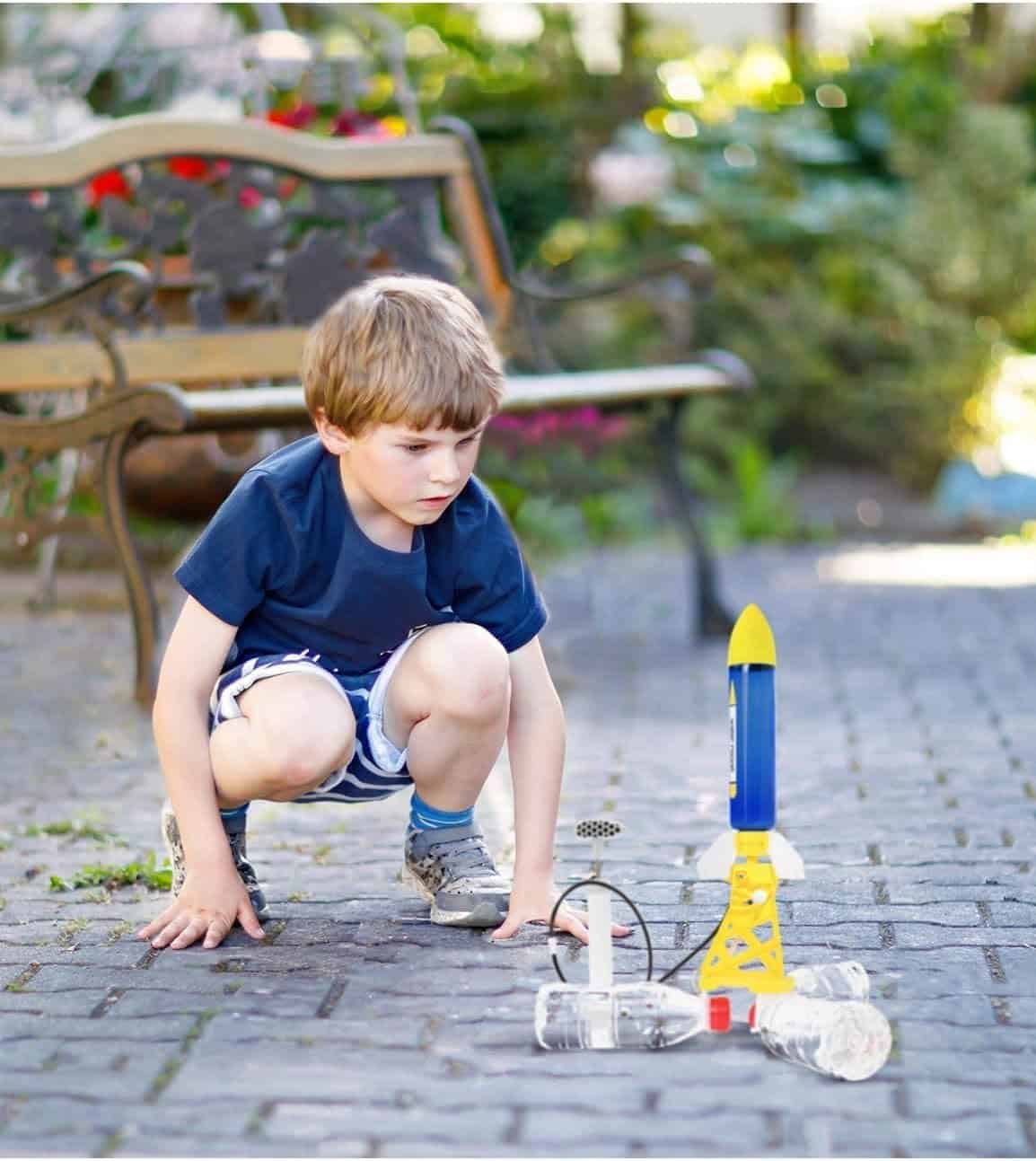 A young boy crouches on a paved surface, watching a toy rocket launcher set up in front of him, with a garden bench and greenery in the background.