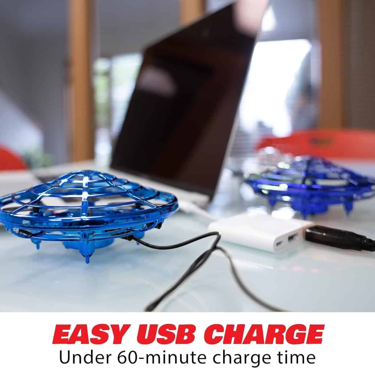 A blue toy drone connected to a power bank via USB cable, charging on a table near a laptop. Text reads: "EASY USB CHARGE Under 60-minute charge time.