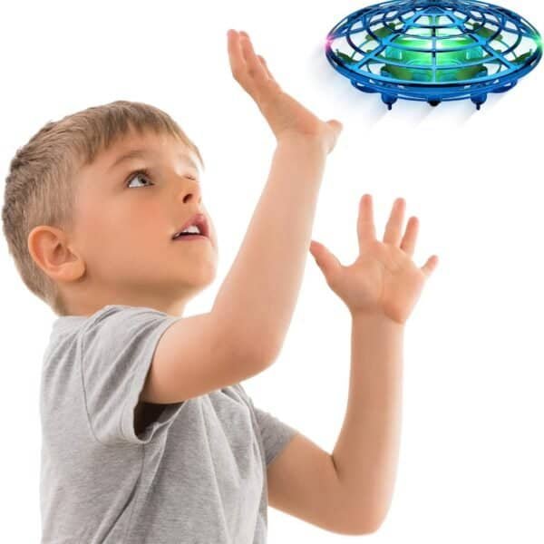 A young boy in a gray shirt raises his hands toward a flying blue drone hovering above him against a white background.