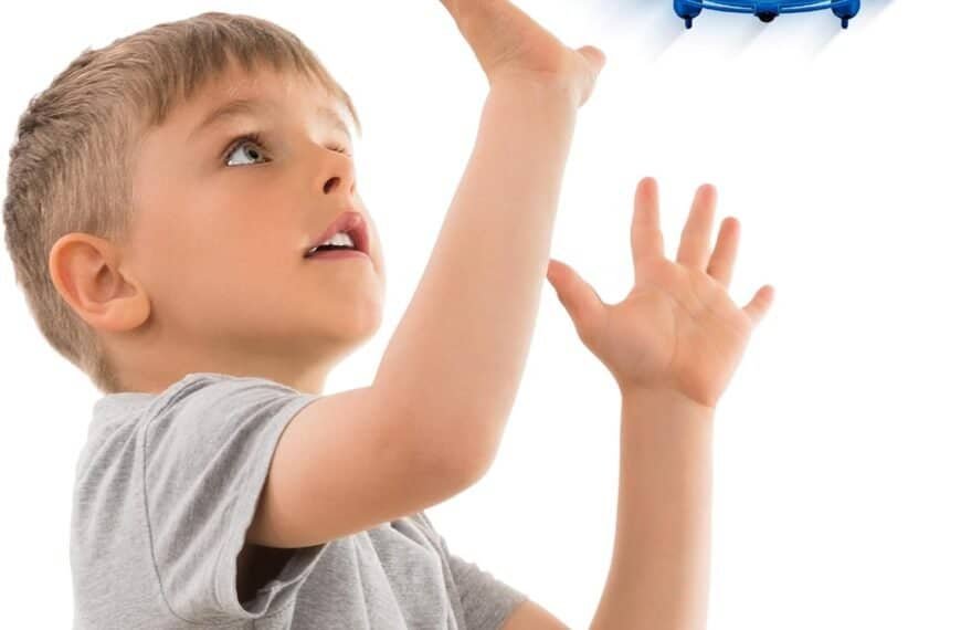 A young boy in a gray shirt raises his hands toward a flying blue drone hovering above him against a white background.
