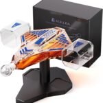 A spaceship-shaped glass whiskey decanter filled with amber liquid, displayed on a black stand, with a branded gift box in the background.