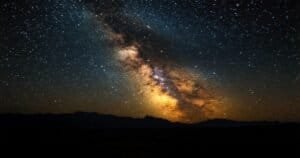 Arco galáctico de la Vía Láctea brillando sobre un paisaje oscuro y accidentado en la noche.
