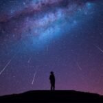 Silhouette of a person standing on a hill at night, gazing up at a star-filled sky with the Milky Way and a mesmerizing meteor shower streaking across the darkness.