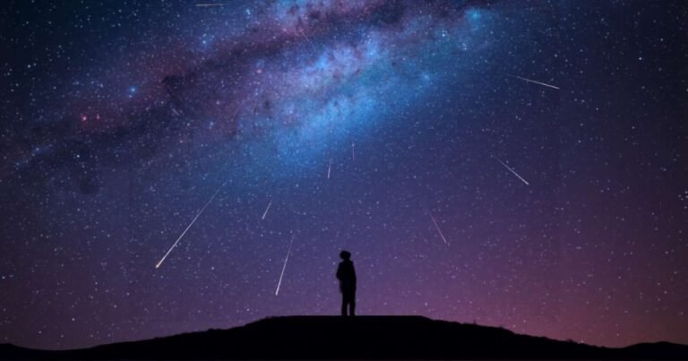 Silhouette of a person standing on a hill at night, gazing up at a star-filled sky with the Milky Way and a mesmerizing meteor shower streaking across the darkness.