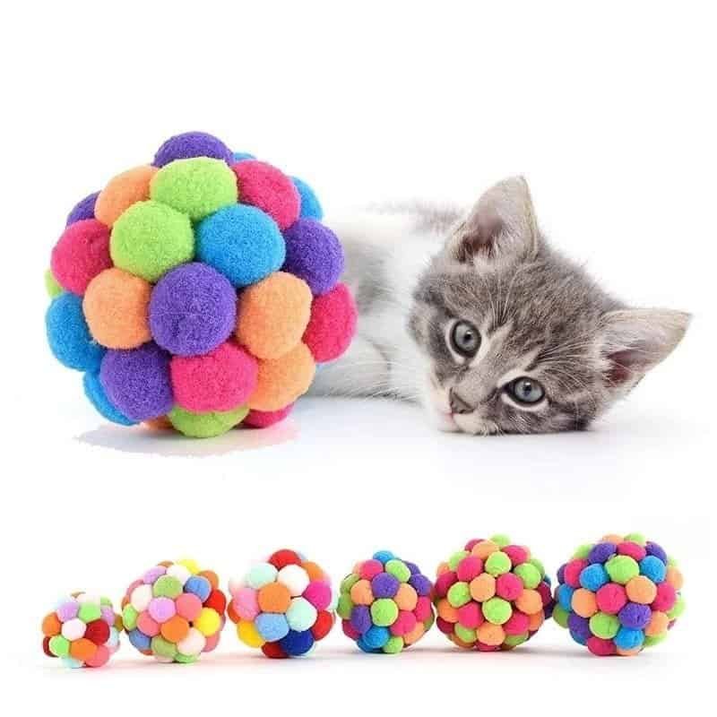 A cat is laying next to a colorful auto-draft pom pom ball.