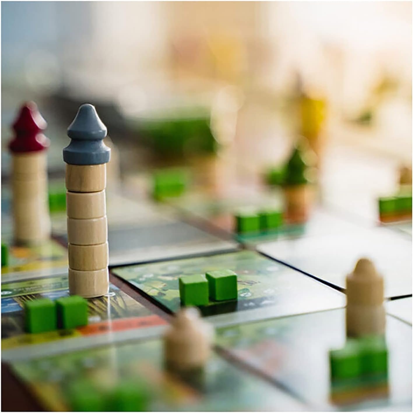Close-up of a tabletop board game featuring a grid of square tiles with small green rectangular pieces and a stack of wooden cylindrical blocks topped with a blue piece resembling a tower.