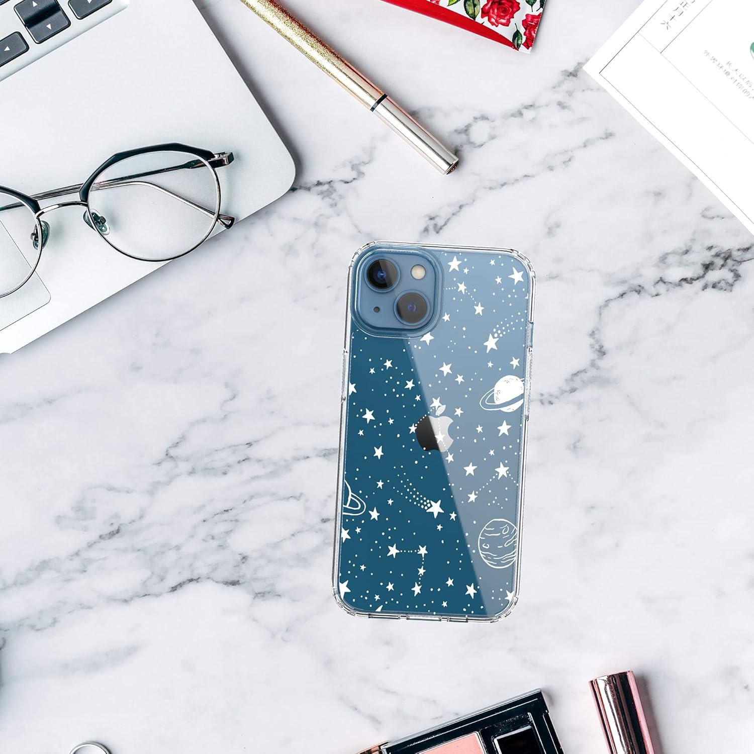 A phone with a space-themed case featuring stars, planets, and rockets is placed on a marble surface next to a pair of glasses, a pen, and a laptop.