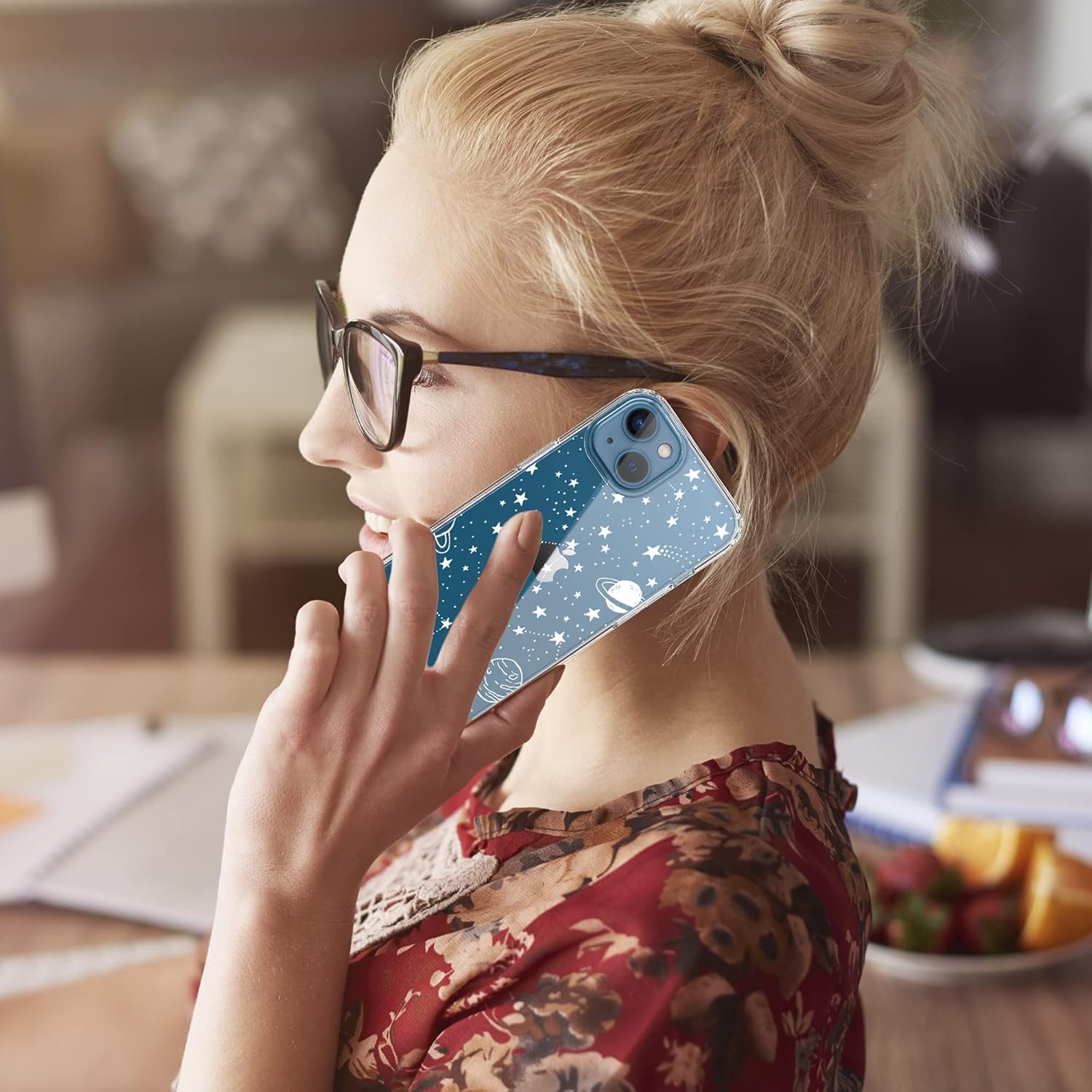 A person wearing glasses and a floral top is holding a phone with a space-themed case to their ear while sitting at a table.