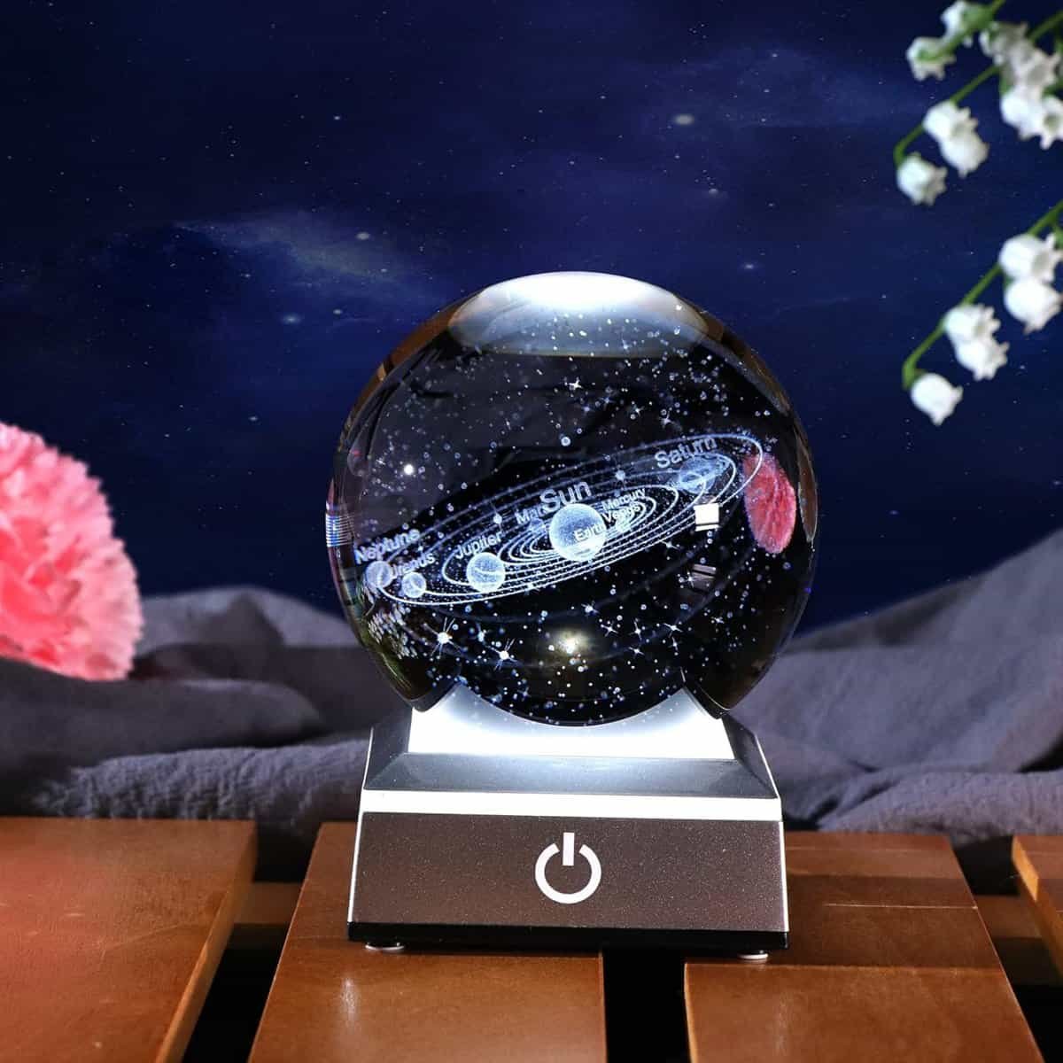 A glass sphere with an engraved solar system model on a lighted base, with a power button, set against a dark background and flowers nearby.
