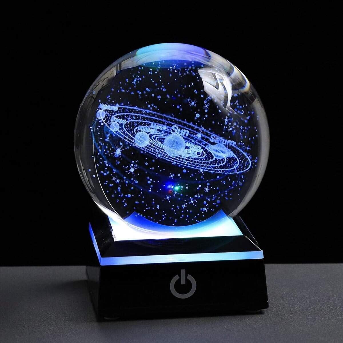 A glass orb displaying a laser-etched solar system on a black base with an illuminated power button symbol.
