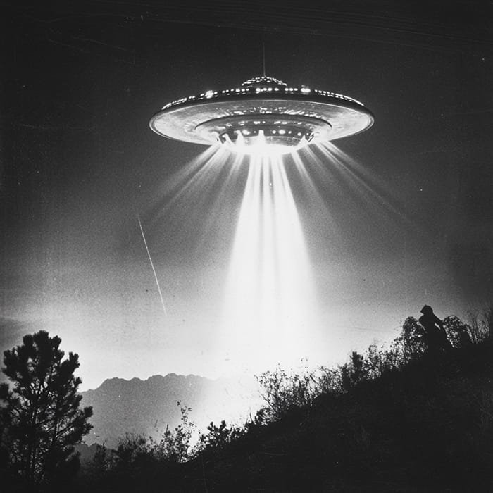 The black and white image captures a mysterious UFO, its bright beams of light illuminating the hillside. Hovering above the trees, it hovers with an otherworldly presence as a lone person gazes upward in awe.