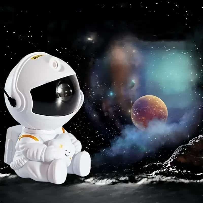 An astronaut figure from the "Your World with the Astronaut Galaxy Projector" perches on a rocky surface, gazing at distant planets and vibrant nebulae.