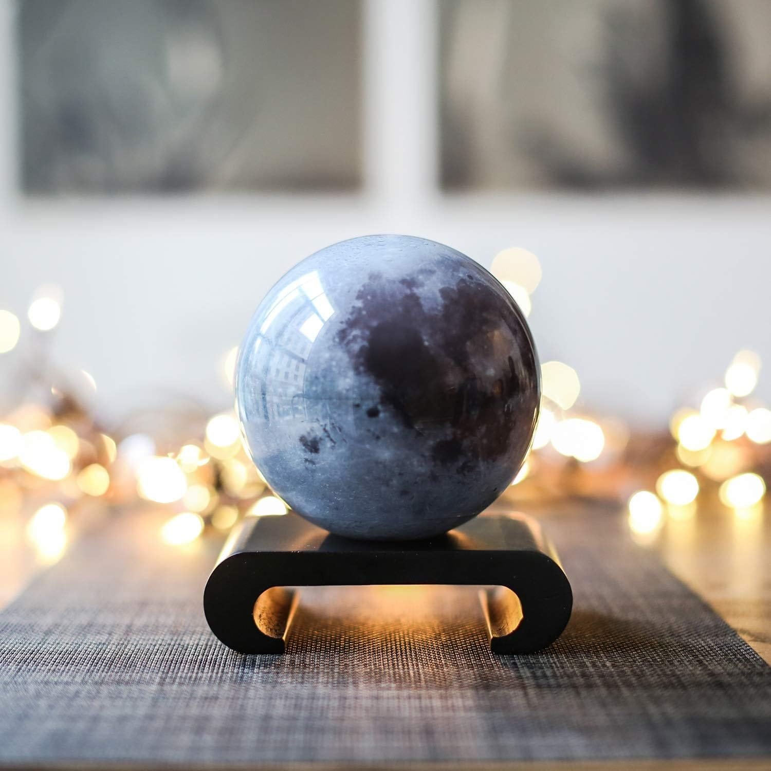 A round, moon-textured sphere sits on a black stand, placed on a gray mat, with soft, blurred lights in the background.